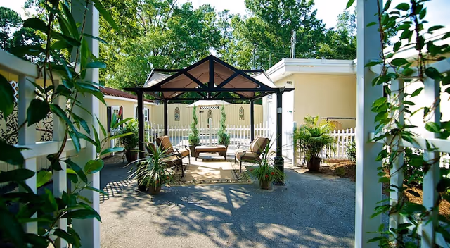Outdoor seating area with a gazebo structure surrounded by potted plants and greenery, located in a courtyard with a white picket fence and light yellow buildings in the background.