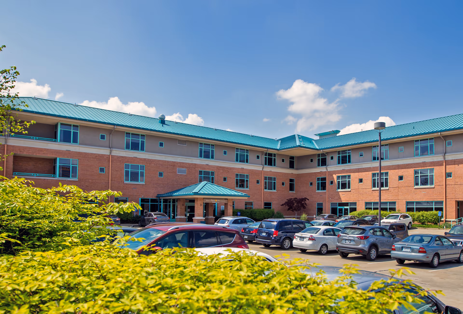 Exterior view of a three-story brick building with teal roof and multiple windows under a blue sky with some clouds. There is a parking lot in front with several parked cars and green bushes in the foreground.