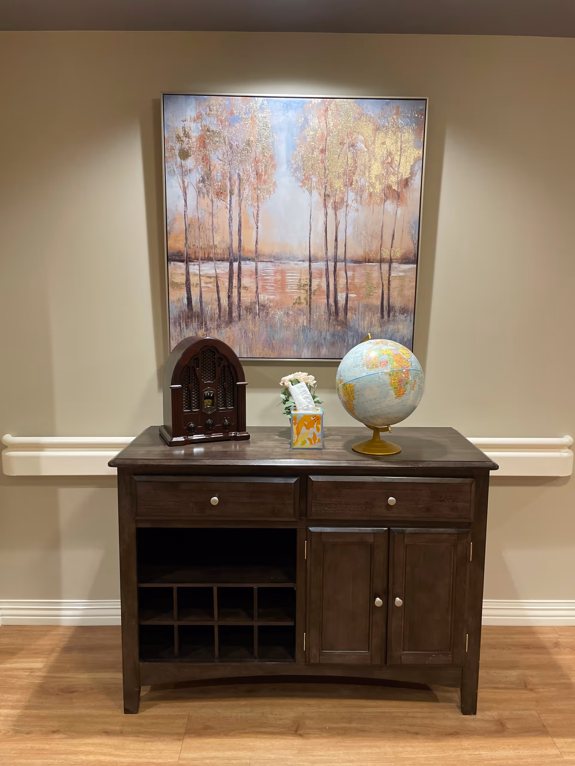 A wooden sideboard with two drawers, two cabinet doors, and open cubby shelves. On top of the sideboard are a vintage-style radio, a tissue box with flowers, and a globe. Above the sideboard hangs a large painting depicting a serene landscape with trees and a body of water.