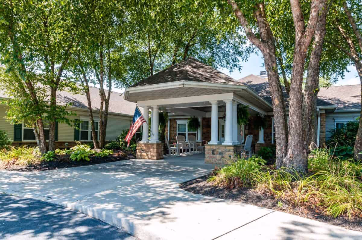 Entrance of a senior living facility with a covered driveway supported by white columns on stone bases. There are several trees and landscaped plants around the driveway, and an American flag is displayed near the entrance. The building has a light-colored exterior with windows and a shingled roof.