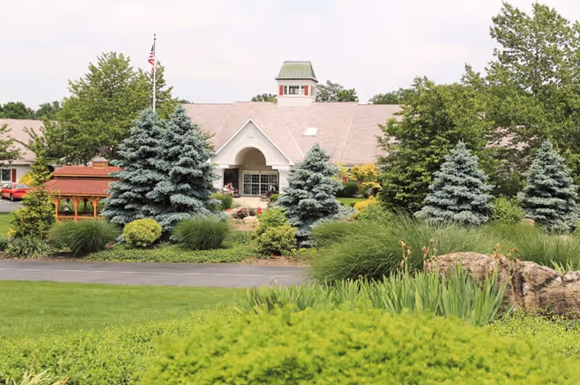 Front exterior view of Oak Leaf Manor North facility with a large building featuring a peaked roof and cupola, surrounded by well-maintained landscaping including evergreen trees, bushes, and a green lawn.