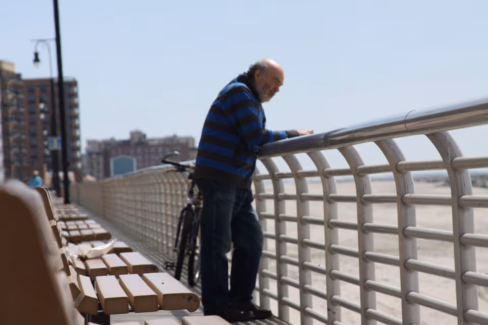 An elderly man wearing a blue and black striped hoodie leans on a metal railing overlooking a beach. There are wooden benches along the walkway and buildings in the background under a clear sky.