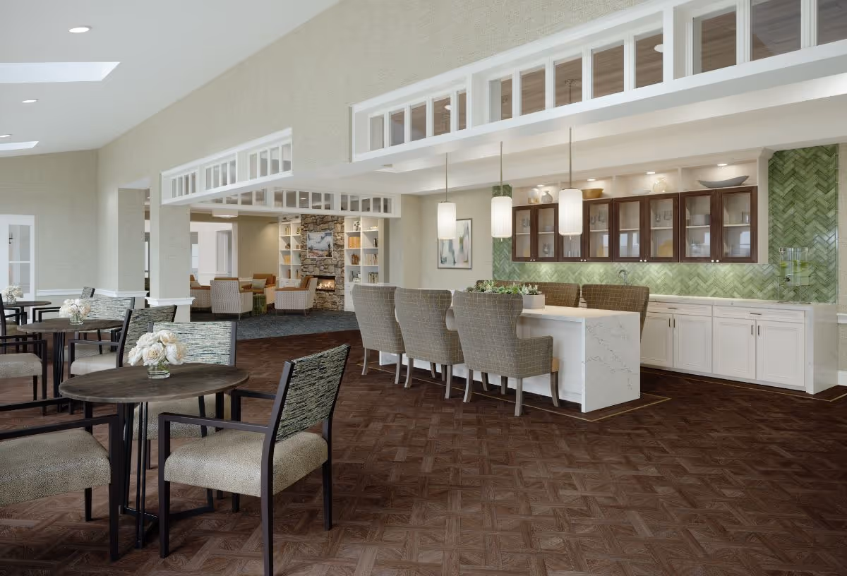 A spacious and modern common area in a senior living facility featuring a seating area with round tables and chairs, a kitchen island with bar stools, pendant lighting, and a green tiled backsplash behind wooden cabinets. In the background, there is a cozy lounge area with armchairs and a stone fireplace.