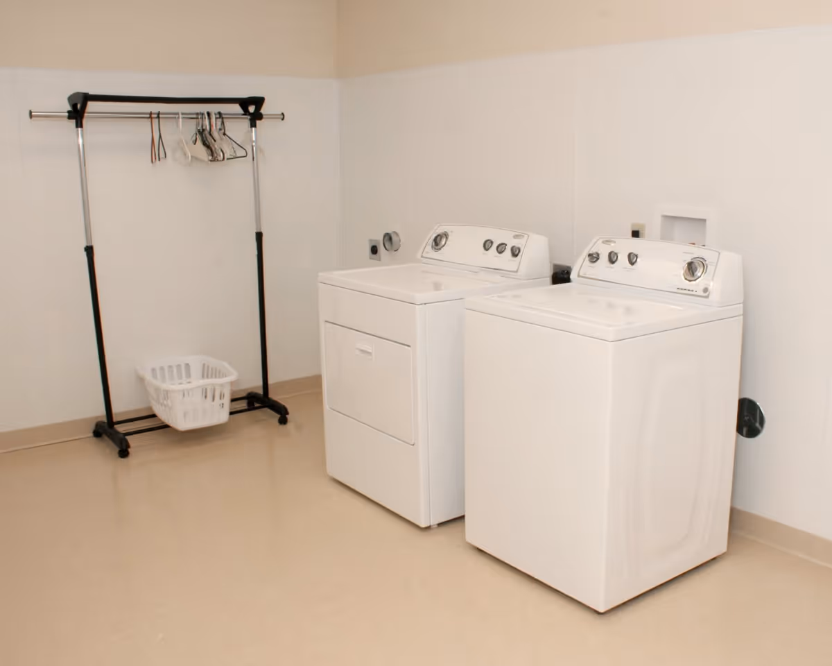 A laundry room with a white washing machine and dryer side by side against a beige wall. To the left, there is a black metal clothing rack with several hangers and a white laundry basket on the floor beneath it. The floor is a light beige color.
