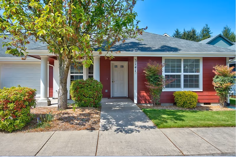 Front exterior view of a single-story red house with white trim, a white front door, large windows with white blinds, a small porch with two white columns, and a concrete walkway leading to the entrance. The yard has green grass, bushes, and a tree with some leaves turning brown.