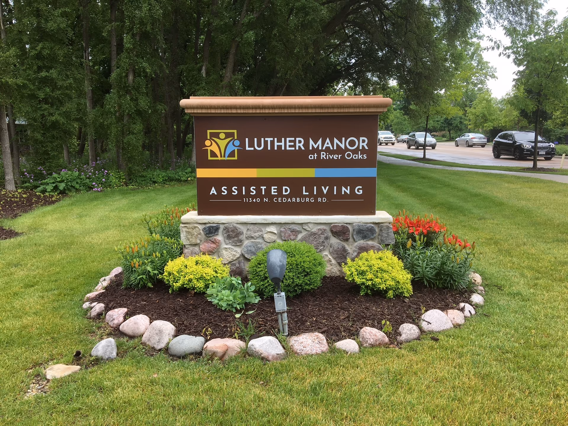 A landscaped sign for Luther Manor at River Oaks Assisted Living, surrounded by green grass, colorful flowers, and small bushes, with a parking lot and several cars visible in the background.