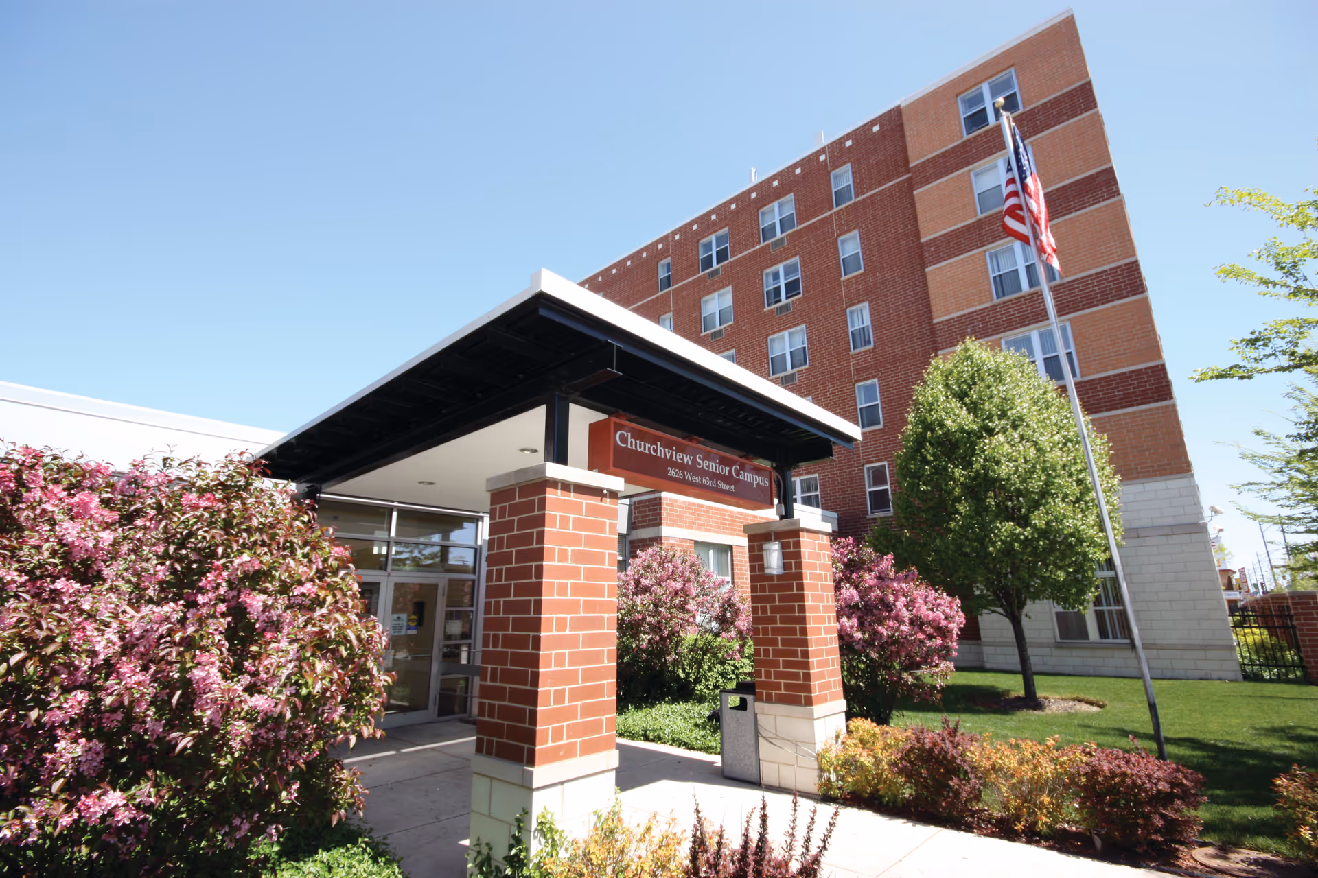 Entrance to Churchview Senior Campus, a multi-story brick building with a covered entryway supported by brick pillars. There are flowering bushes and trees around the entrance, and an American flag on a flagpole nearby under a clear blue sky.