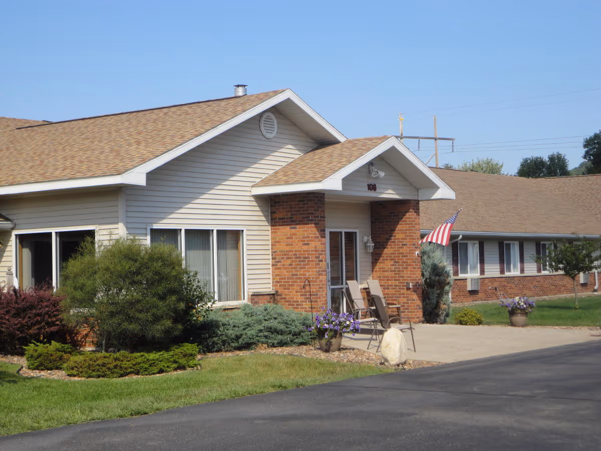 Exterior view of a single-story senior living facility building with beige siding and brick accents. There is a small covered entrance with two chairs and an American flag mounted on the wall. The building is surrounded by well-maintained landscaping including bushes, flowers, and a green lawn. The sky is clear and blue.