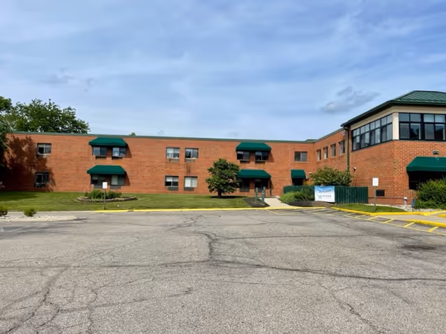 Exterior view of a two-story brick senior living facility with green awnings over some windows and entrances. There is a parking lot in the foreground and a small tree and shrubs near the building.