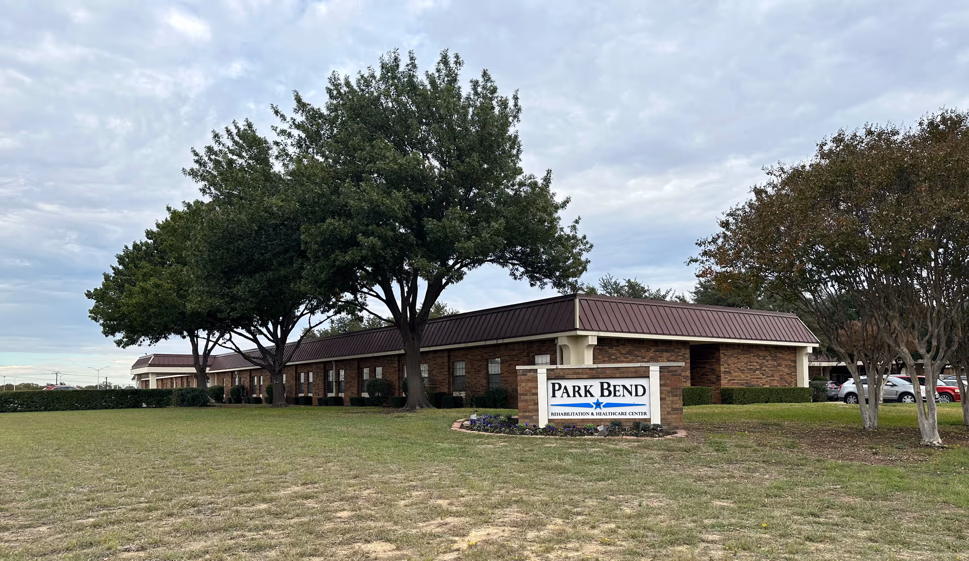 Exterior view of Park Bend Rehabilitation & Healthcare Center, a single-story brick building with a brown roof, surrounded by trees and a grassy lawn. A sign in front displays the facility's name.