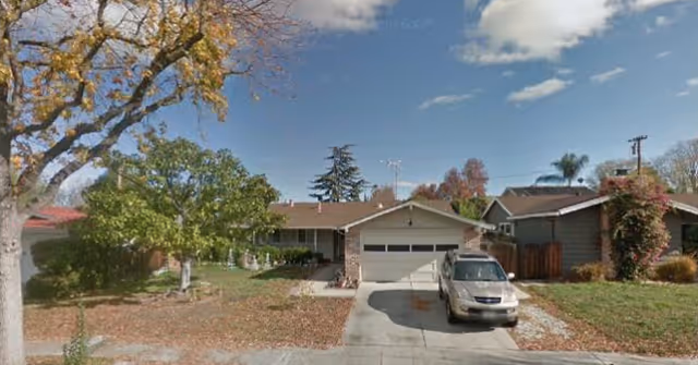 Front exterior view of a single-story residential home with a driveway, a parked silver SUV, a large tree on the left, and a clear blue sky with some clouds.