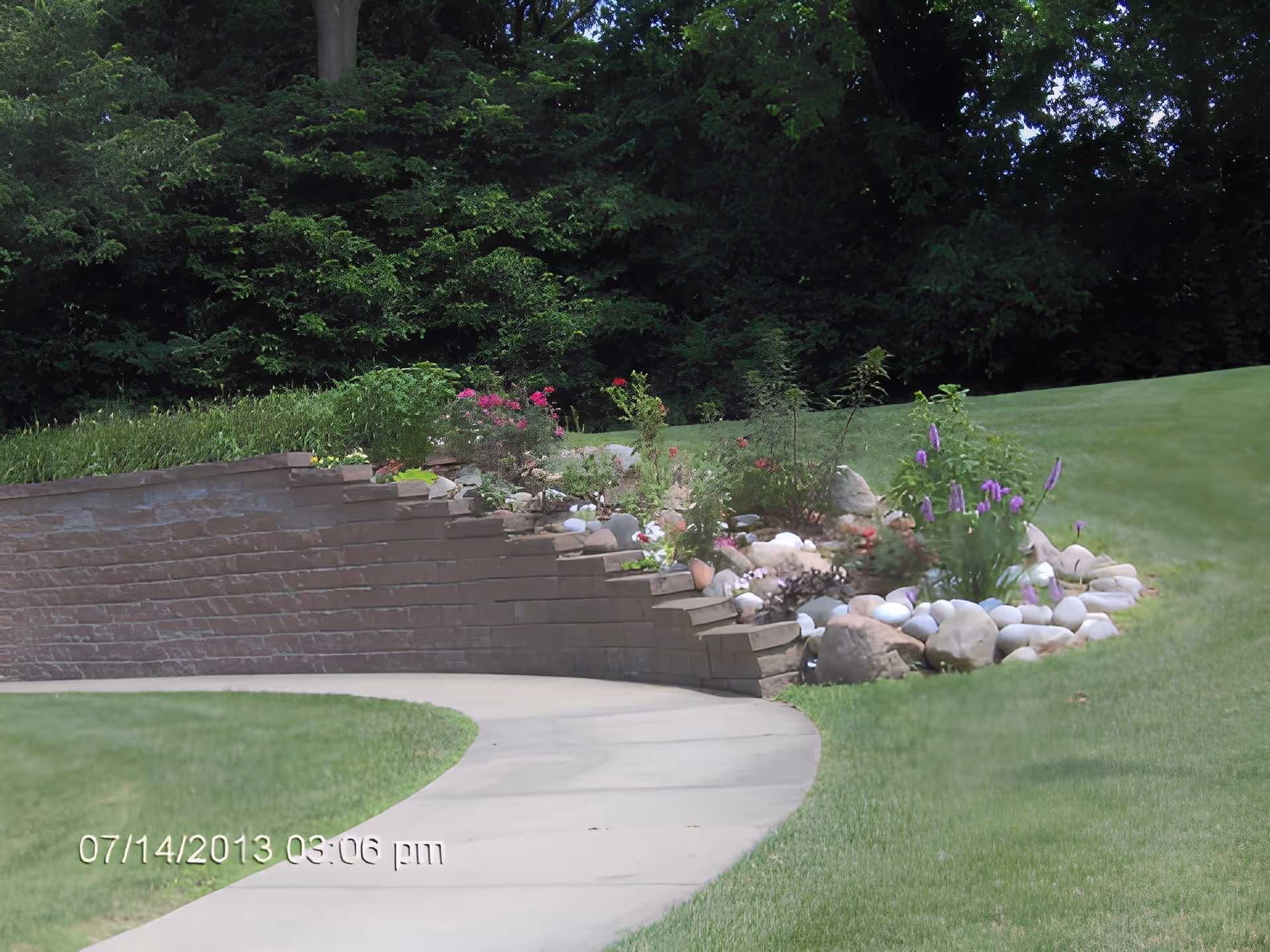 A curved concrete pathway bordered by a low brick retaining wall with various flowering plants and rocks. The background features dense green trees and a well-maintained grassy area.
