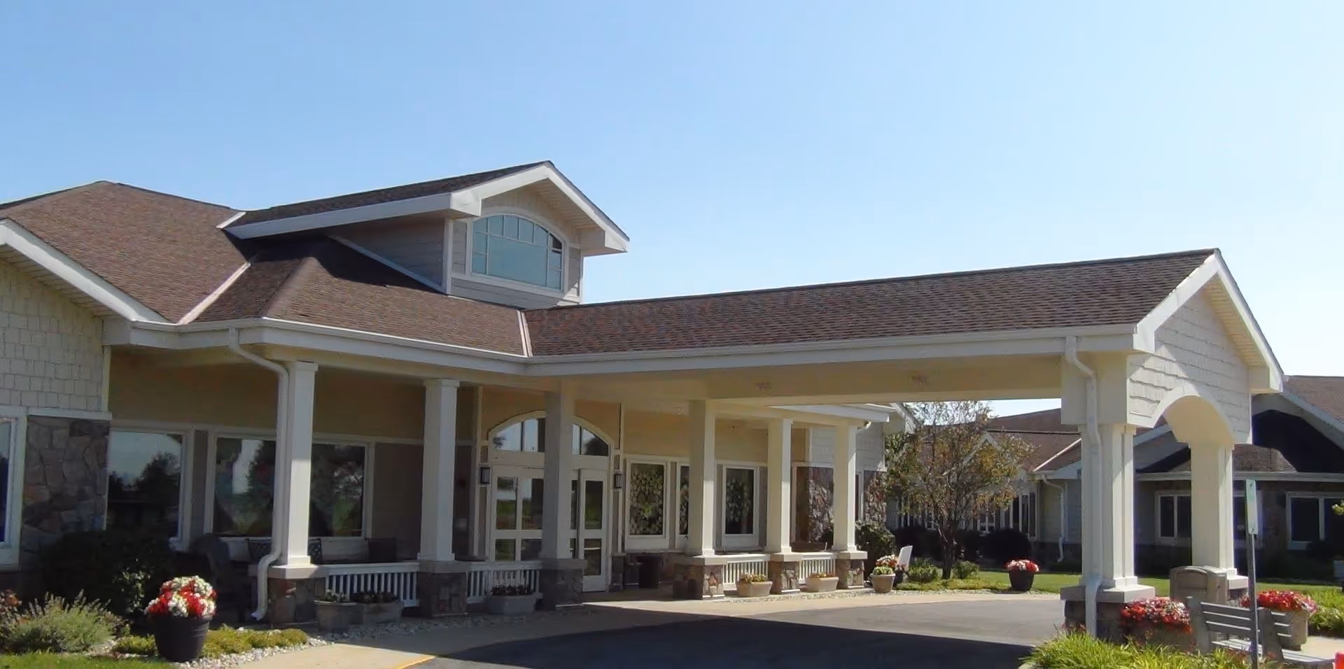 Exterior view of Lakeland Health Care Center showing the main entrance with a covered drop-off area supported by white columns. The building has a beige facade with stone accents and a brown shingled roof under a clear blue sky.