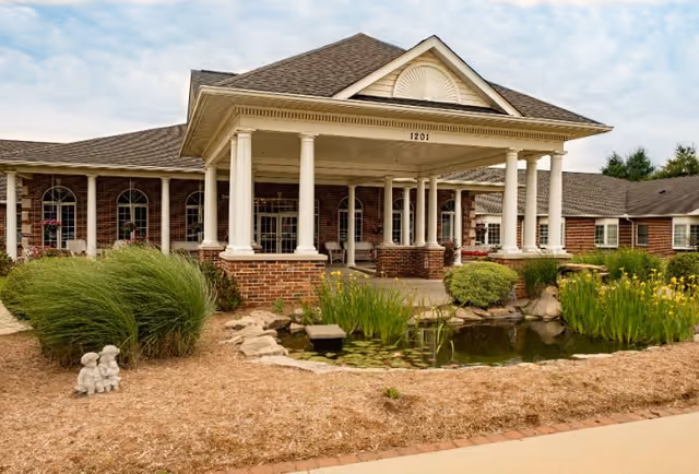 Exterior view of Charter Senior Living of Shiloh showing a brick building with white columns supporting a covered entrance. The building number 1201 is visible above the entrance. In front of the building, there is a landscaped area with green bushes, tall grasses, yellow flowers, a small pond with lily pads, and a small stone statue of two children sitting together.