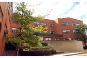 Exterior view of a multi-story brick building with several windows, surrounded by trees and shrubs under a partly cloudy sky.