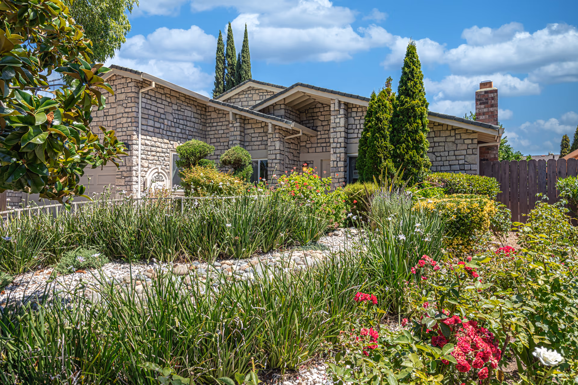 Stone exterior of a single-story building with a well-maintained garden featuring various green shrubs, tall trees, and blooming red and white flowers under a partly cloudy blue sky.