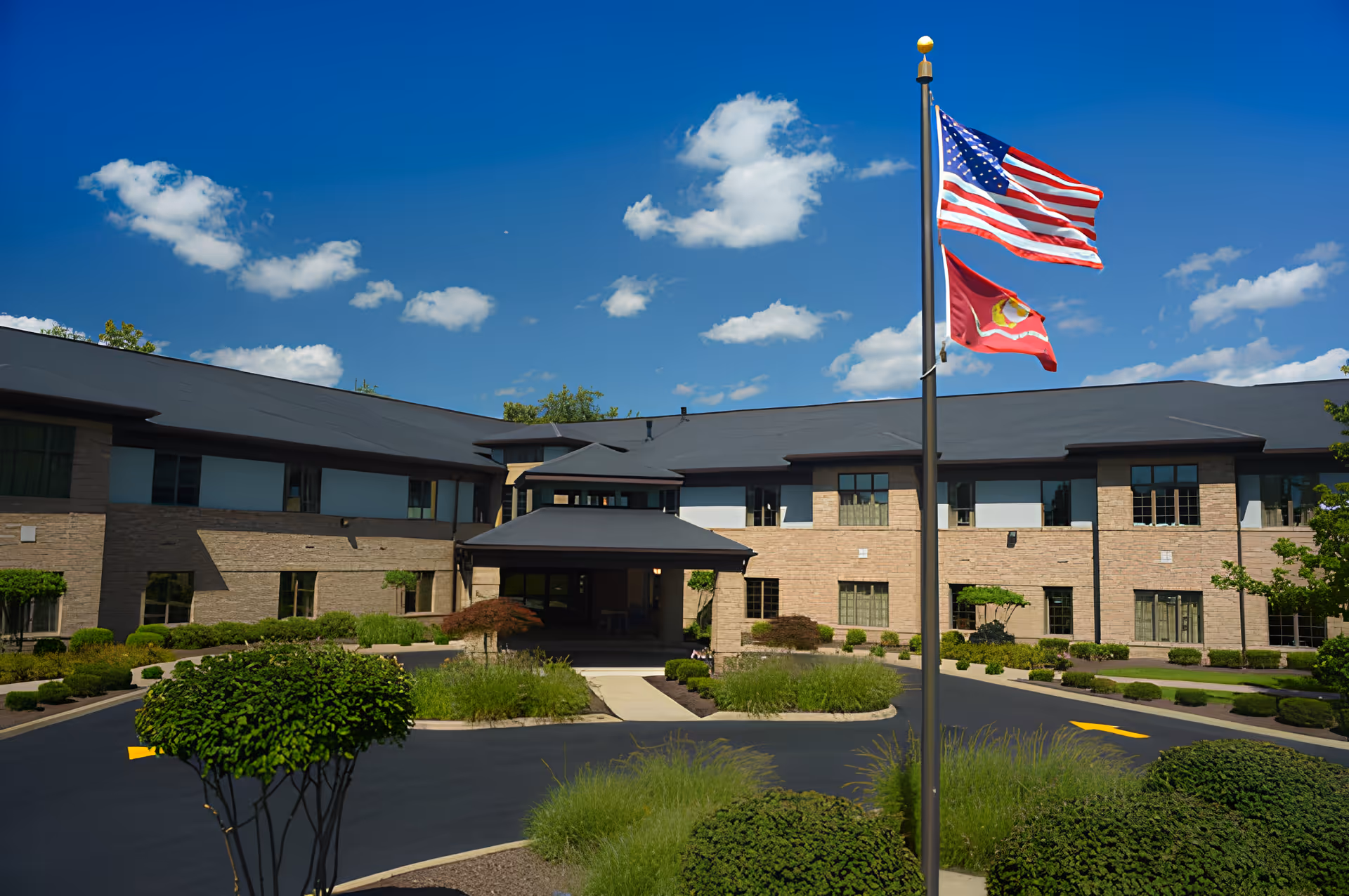 Exterior view of Park Village Health Care building with a clear blue sky and scattered clouds. The building is two stories with a covered entrance, surrounded by neatly trimmed bushes and landscaped greenery. Two flags, including the American flag, are flying on a flagpole in front of the building.