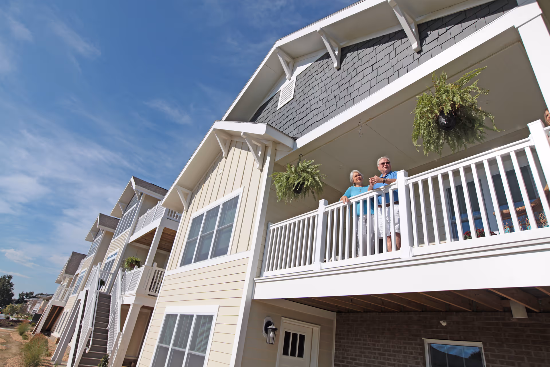 An elderly couple standing on a white balcony of a multi-story residential building with hanging plants, under a blue sky with some clouds.