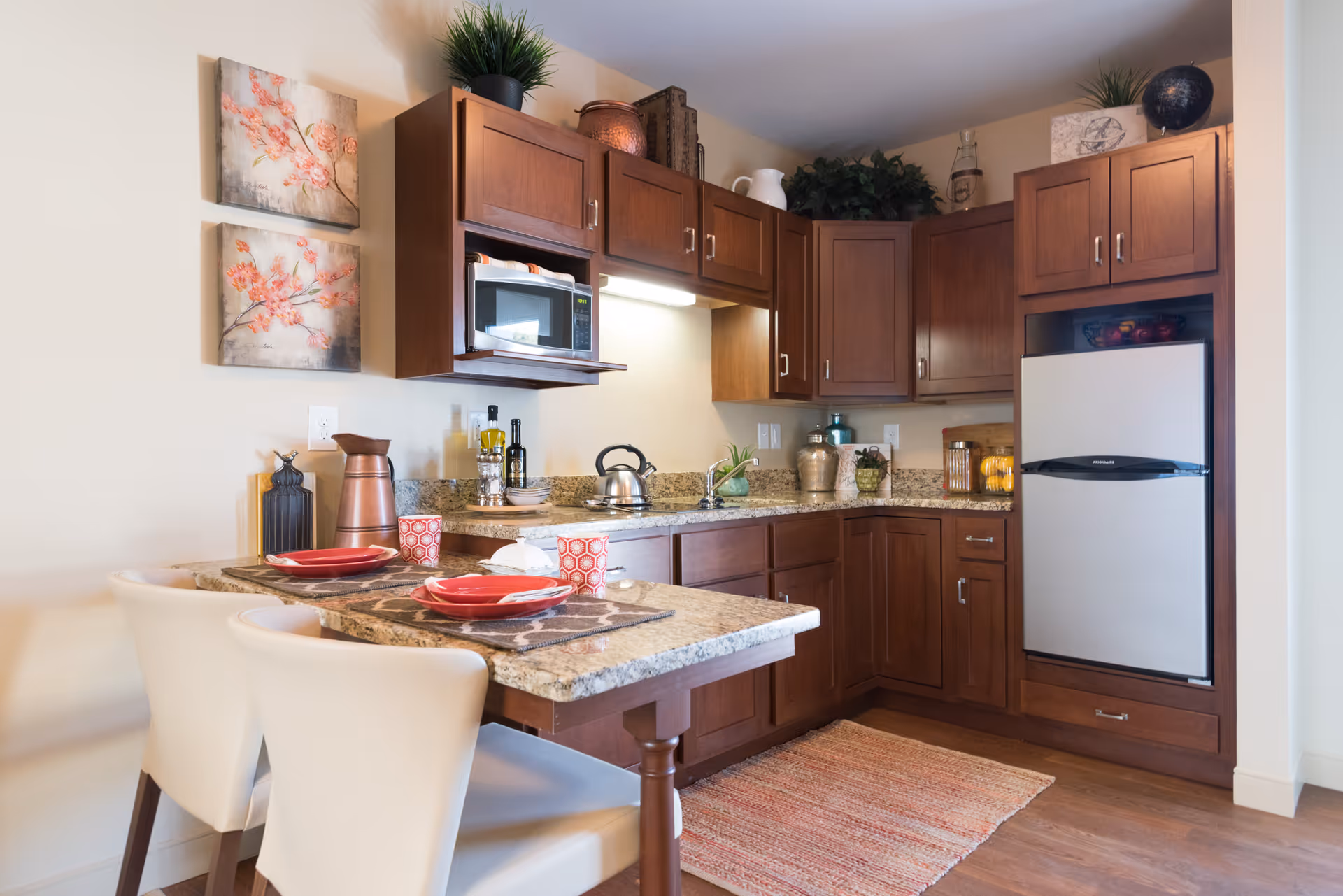 A modern kitchen with wooden cabinets, granite countertops, and a small dining area with two white chairs and place settings. The kitchen includes a microwave, refrigerator, kettle, and decorative items such as plants and artwork on the wall.