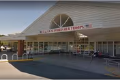 Exterior view of the entrance to Long Island State Veterans Home with a covered drop-off area. A banner above the entrance reads 'LONG ISLAND SUPPORTS OUR TROOPS!'. The building has large windows and a semi-circular window above the entrance. There are bike racks and a wheelchair ramp visible near the entrance.