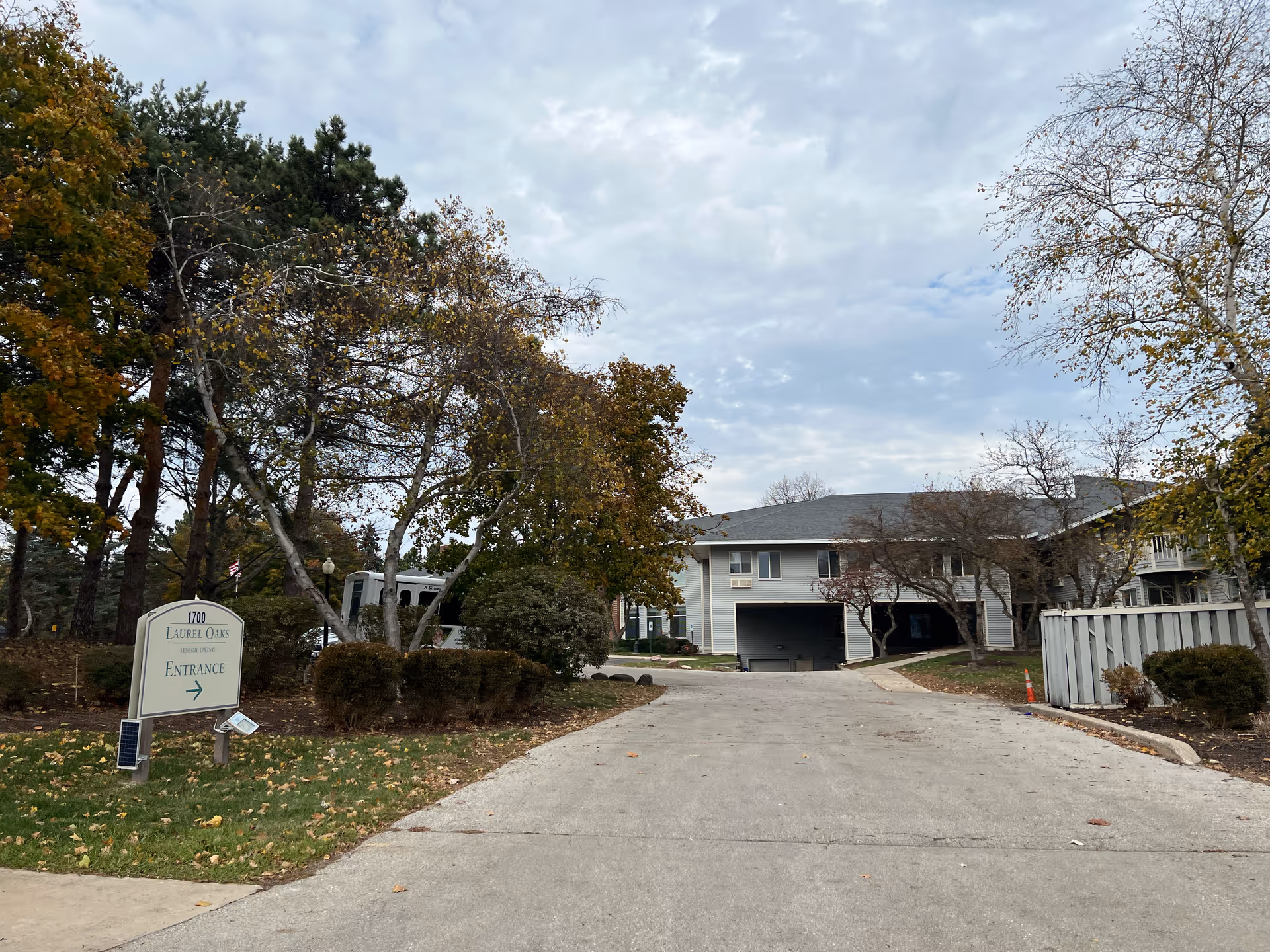 Driveway leading to a two-story senior living facility building with trees and bushes on both sides. A sign on the left reads '1700 Laurel Oaks Senior Living Entrance' with an arrow pointing right. The sky is cloudy and the trees have autumn foliage.