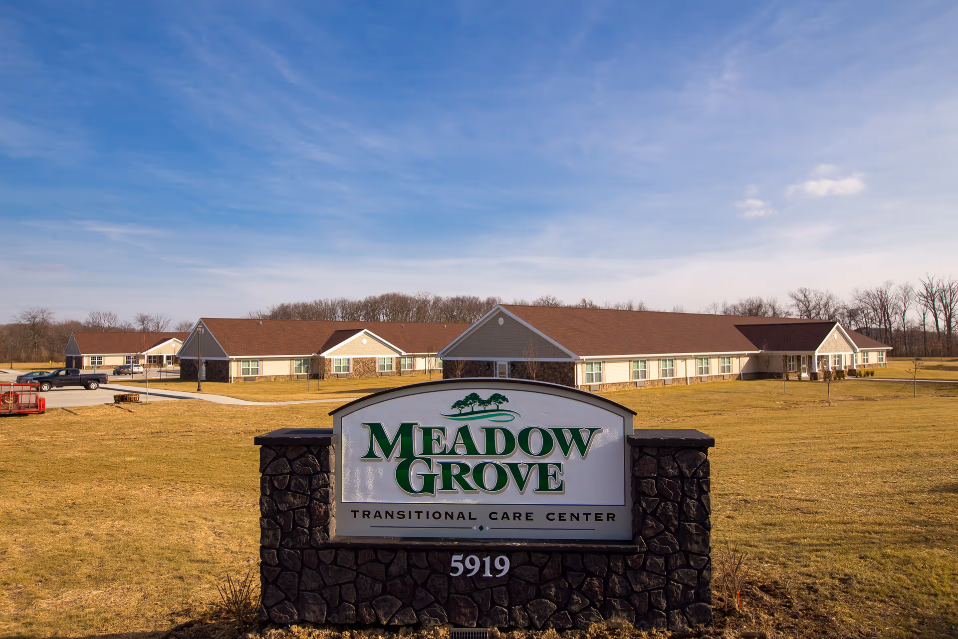 View of Meadow Grove Transitional Care Center building with a large stone sign in the foreground displaying the facility name and address number 5919. The building is a single-story structure with a brown roof and beige siding, surrounded by a grassy area under a partly cloudy blue sky.