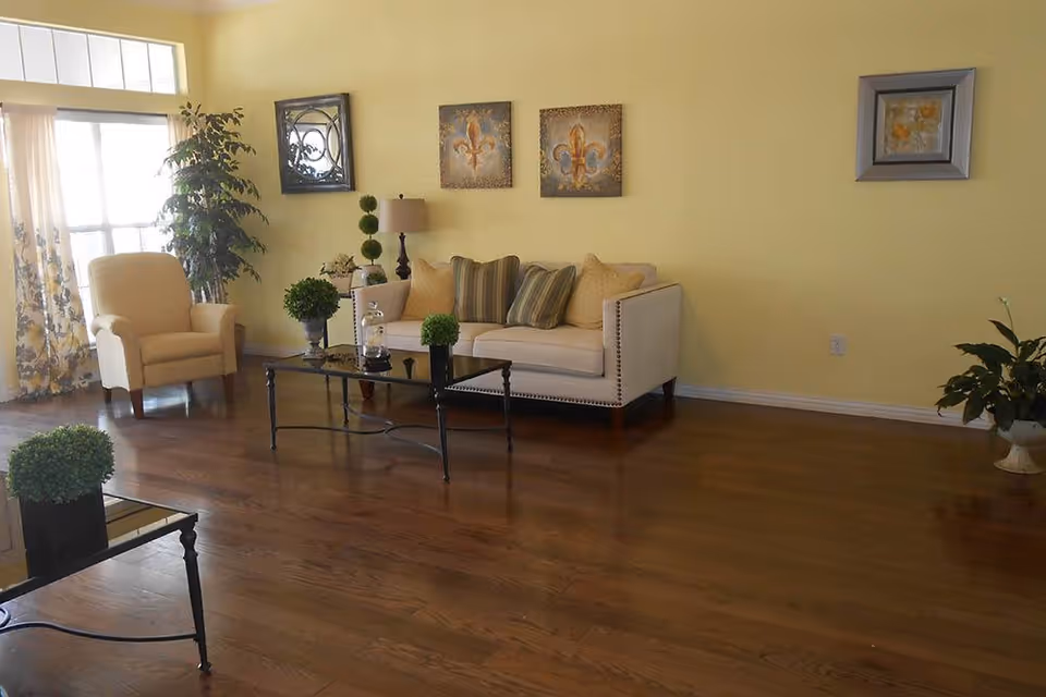 A cozy living room with yellow walls, a beige sofa with multiple cushions, a beige armchair, a glass coffee table with decorative plants, a floor lamp, and framed artwork on the walls. The room has wooden flooring and a large window with sheer curtains allowing natural light to enter.