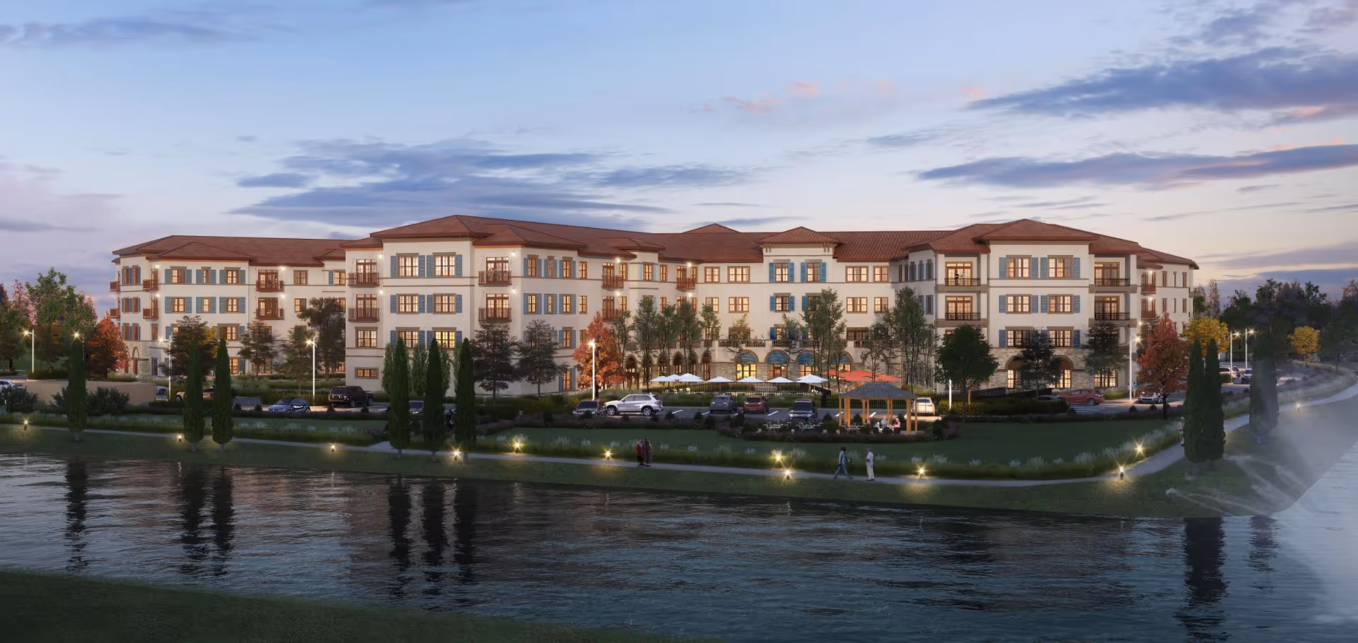 Exterior view of a large, multi-story senior living facility named Grand Living At Tuscan Lakes, featuring a Mediterranean-style design with white walls and red-tiled roofs. The building is surrounded by landscaped greenery, trees, a walking path along a water body, parked cars, and outdoor seating areas under umbrellas. The scene is set during dusk with lights illuminating the grounds.