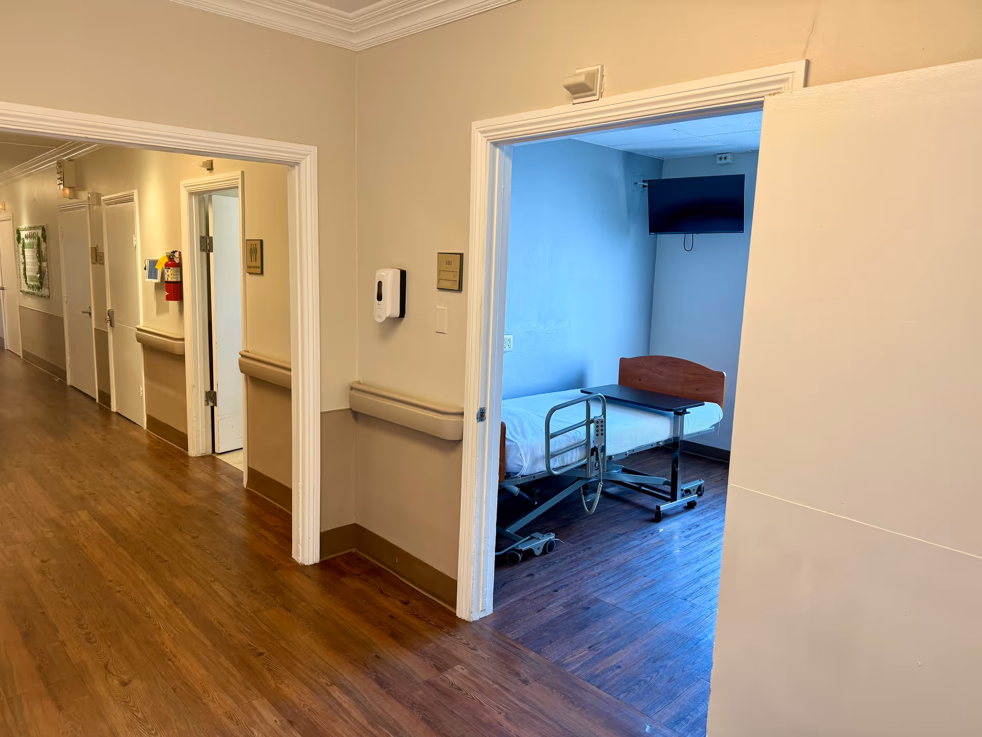 View down a hallway in a senior living facility with wooden flooring and beige walls. Several doors line the hallway, one of which is open revealing a small room with a hospital bed, an overbed table, and a wall-mounted TV.