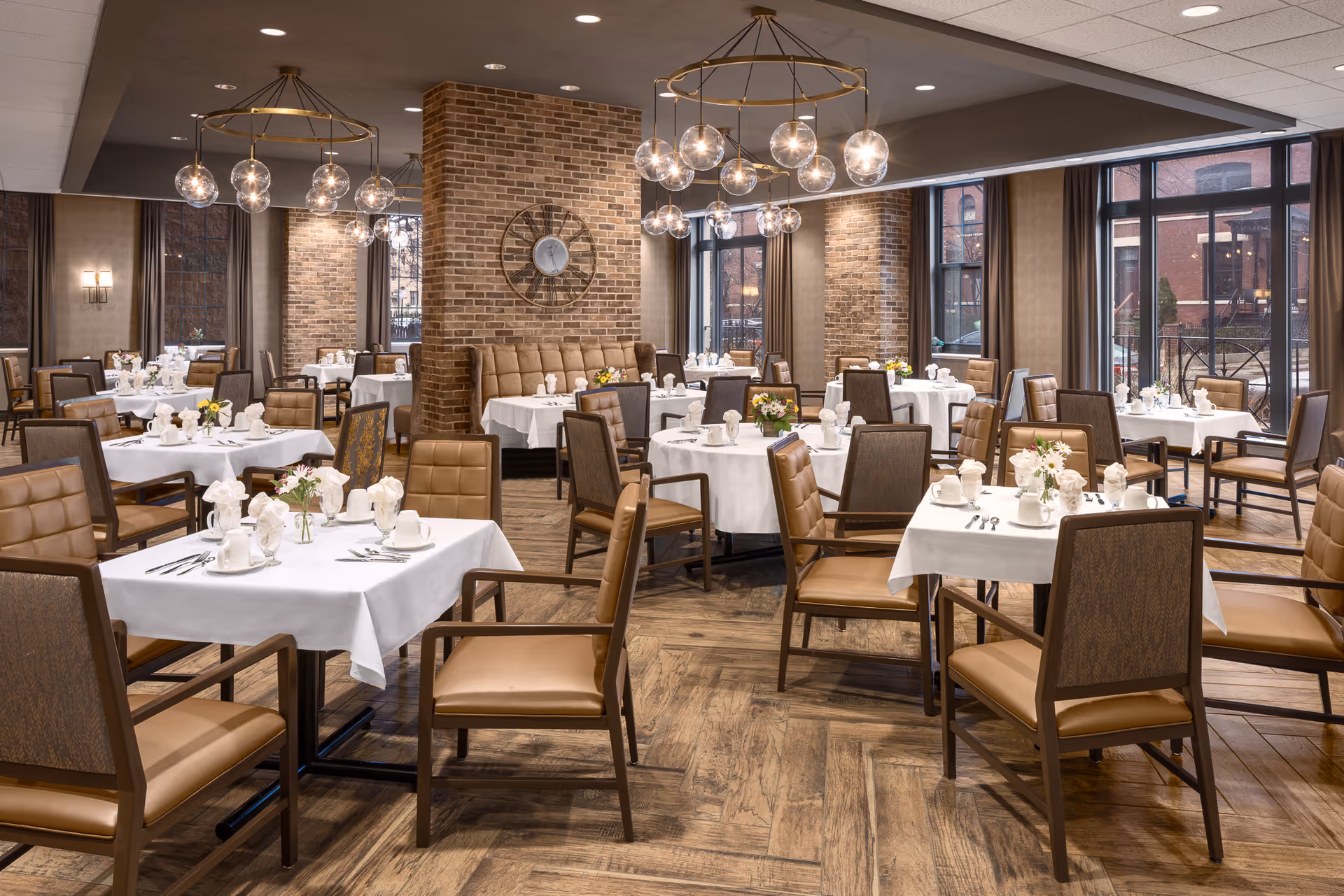 A spacious, well-lit dining room with multiple tables covered in white tablecloths, each set with napkins, cups, and silverware. The room features brown leather chairs, brick accent walls, large windows letting in natural light, and modern spherical chandeliers hanging from the ceiling.
