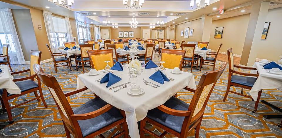 A spacious dining room with multiple tables covered in white tablecloths, each set with blue folded napkins, white cups, glasses, and silverware. The room has patterned carpet, wooden chairs with blue and yellow cushions, chandeliers hanging from the ceiling, and large windows letting in natural light.