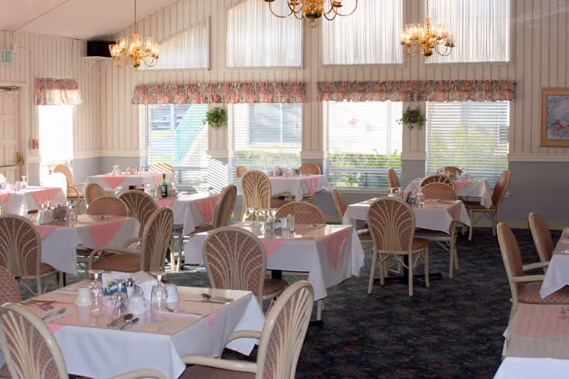 A bright dining room with multiple tables covered in white tablecloths and pink napkins. Each table is set with glasses, silverware, and condiments. The room features large windows with blinds and floral valances, chandeliers hanging from the ceiling, and cushioned chairs with a woven pattern. The carpet is dark with a floral design.