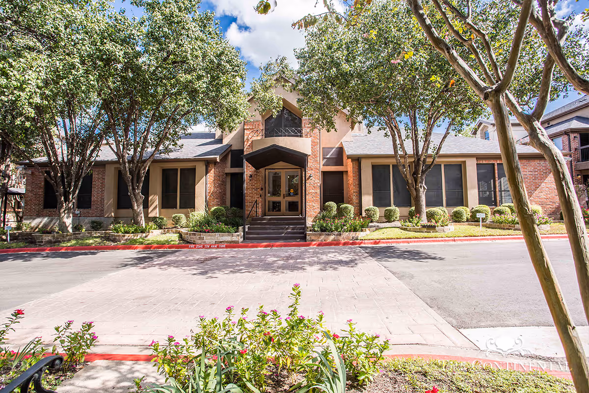 Front exterior view of The Continental Retirement Community building with a paved driveway, landscaped garden with flowers and bushes, and several trees providing shade under a partly cloudy sky.