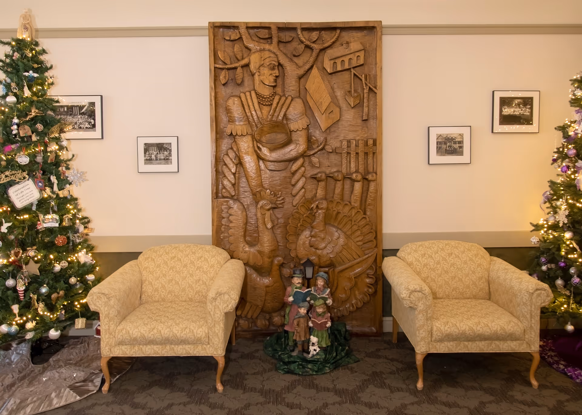 Two beige upholstered armchairs are positioned on either side of a large wooden carved panel depicting a person with birds and a tree. In front of the panel is a figurine of four carolers with a small dog. On both sides of the chairs are decorated Christmas trees with lights and ornaments. The wall behind has four framed black and white photographs.