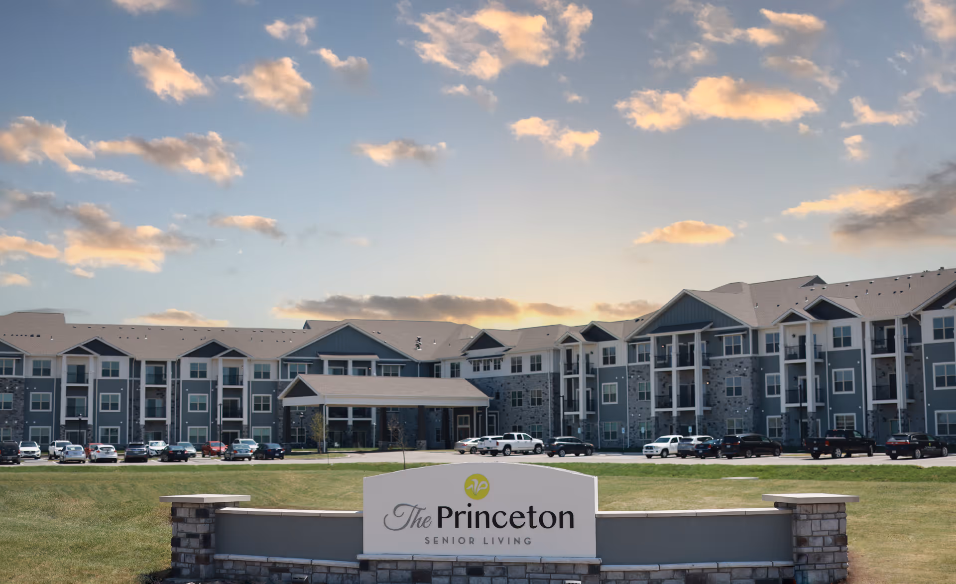 Exterior view of The Princeton Senior Living facility, a large multi-story building with a stone and siding facade, several parked cars in front, and a sign in the foreground displaying the facility's name. The sky is partly cloudy with a warm light from the setting or rising sun.