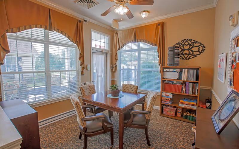 A cozy room with a square wooden table surrounded by four upholstered chairs. The room has large windows with blinds and mustard-colored valances, allowing natural light to fill the space. There is a bookshelf filled with books and games, a decorative wall piece, and a small plant centerpiece on the table. The carpet has a patterned design, and the walls are painted a warm beige color.