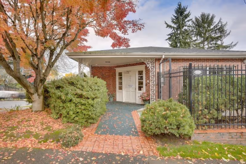 Entrance to a brick building with a covered porch, white double doors, and decorative white metal supports. The walkway is lined with bushes and trees with autumn-colored leaves scattered on the ground.