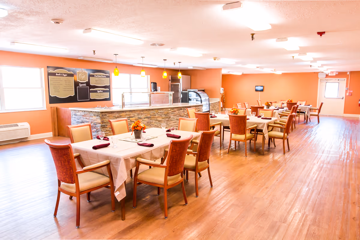 A bright and spacious dining room with several tables covered in white tablecloths, each set with glasses and napkins. The chairs around the tables have wooden frames and cushioned seats. The walls are painted a warm orange color, and there is a serving counter with a stone facade and a menu board on the wall behind it. The room has large windows letting in natural light and a wooden floor.