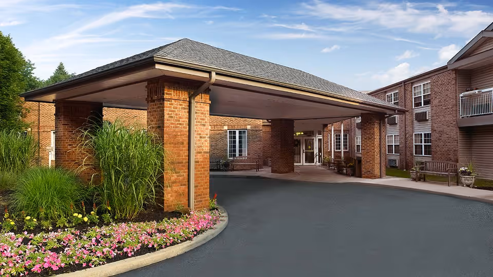 Entrance of a senior living facility with a covered drop-off area supported by brick pillars, surrounded by landscaped flower beds and greenery. The building is made of brick with multiple windows and benches near the entrance.