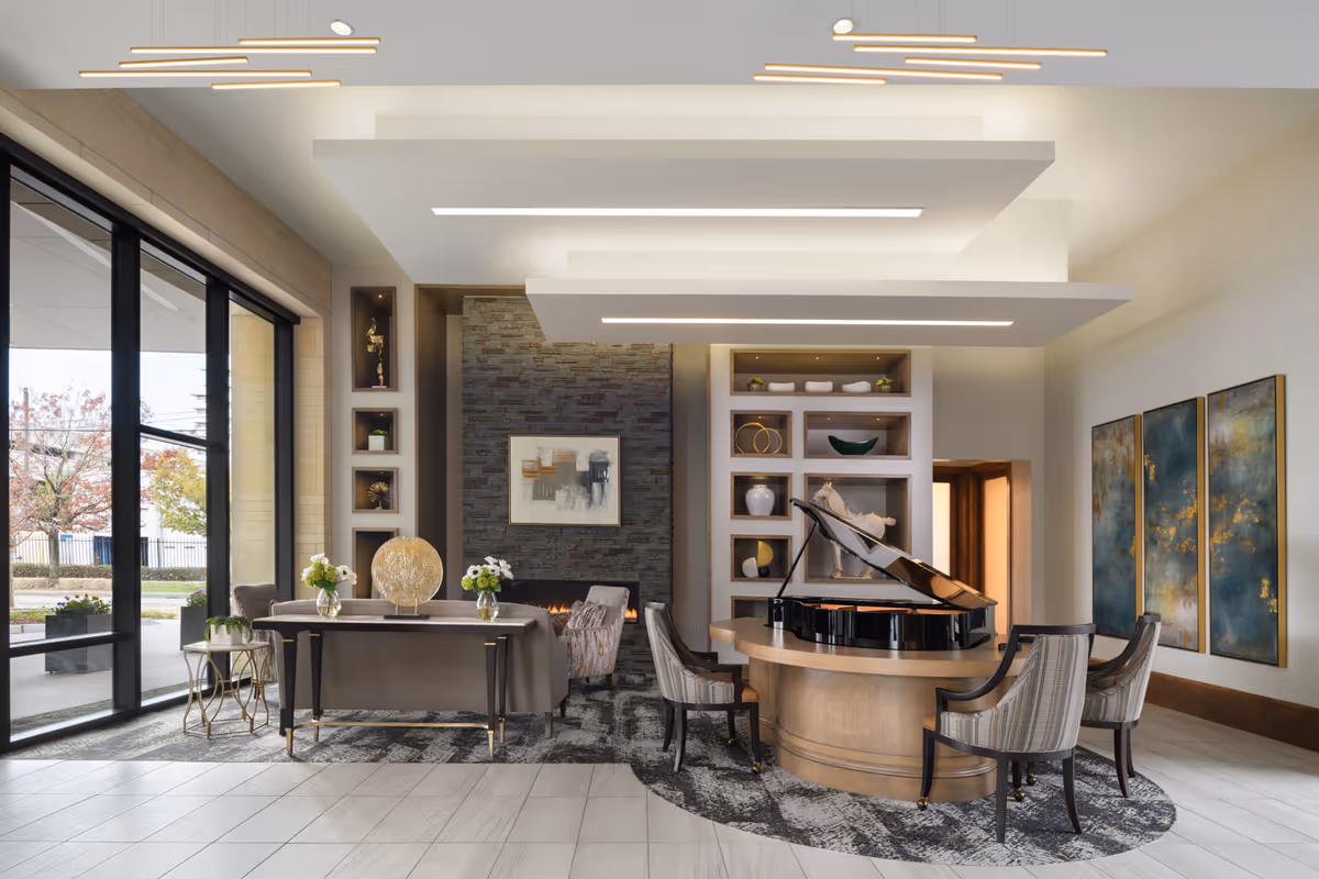 A modern senior living facility common area with a grand piano on a round wooden table surrounded by four upholstered chairs. To the left, there is a seating area with a sofa, armchairs, and a fireplace with a stone wall backdrop. Large windows allow natural light to fill the room, and decorative shelves and artwork adorn the walls.