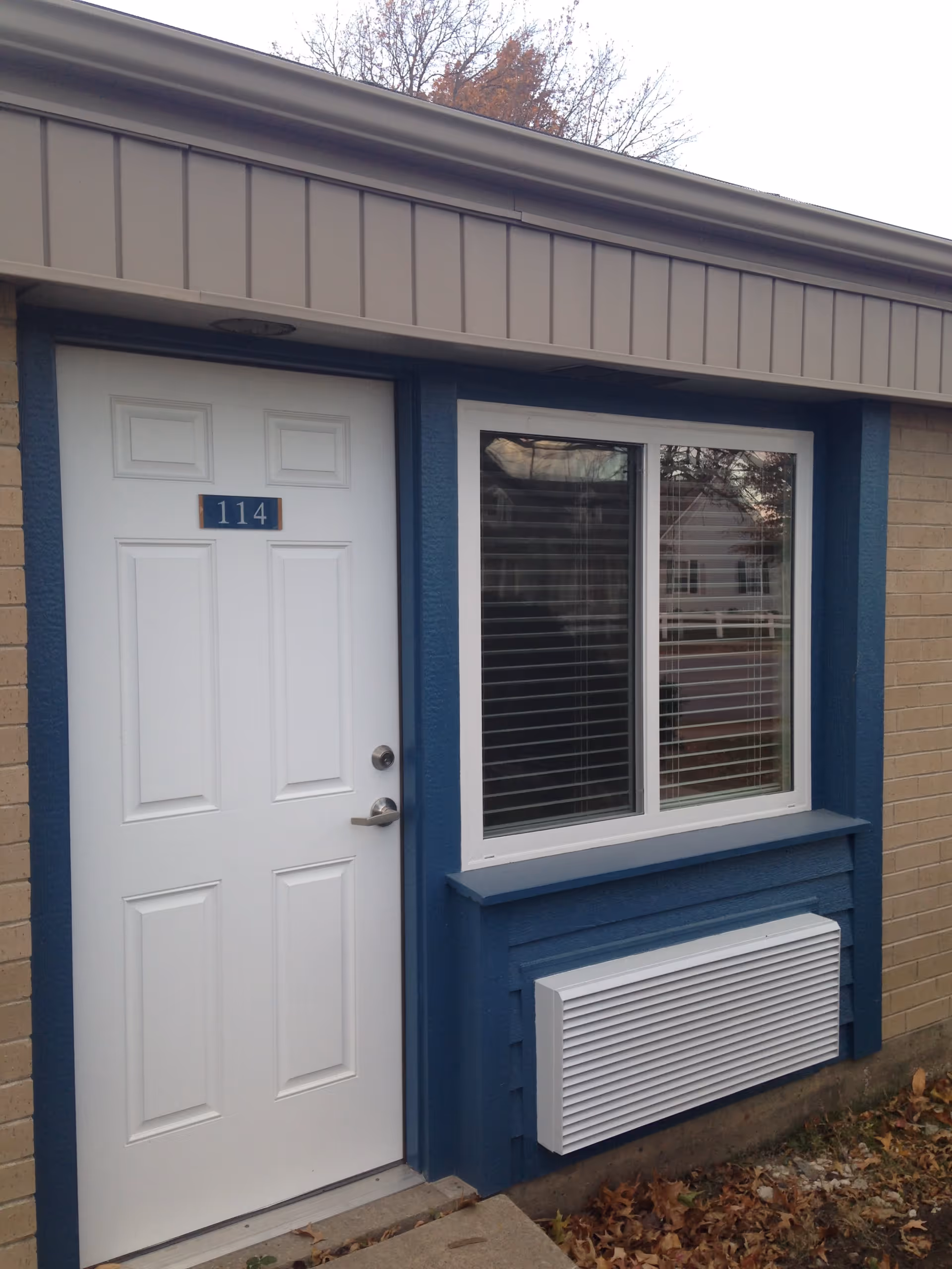 Exterior view of a building entrance with a white door labeled 114 and a window with closed blinds next to it. The building has beige brick walls and blue trim around the door and window. There is an air conditioning unit below the window and some fallen leaves on the ground.