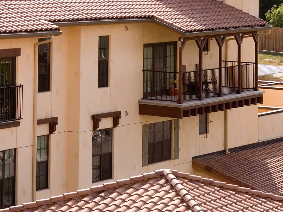 Exterior view of a beige stucco building with a red tile roof and a balcony with wooden beams and black metal railing. The balcony has two chairs and a small table with a potted plant. Several windows are visible on the building's facade.
