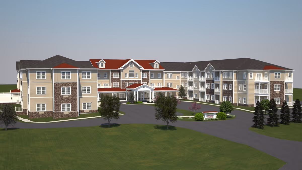 Exterior view of a large three-story senior living facility building with beige and stone facade, red and gray roofs, multiple windows, balconies, and a covered entrance. The building is surrounded by a paved driveway, green lawn, and several trees.