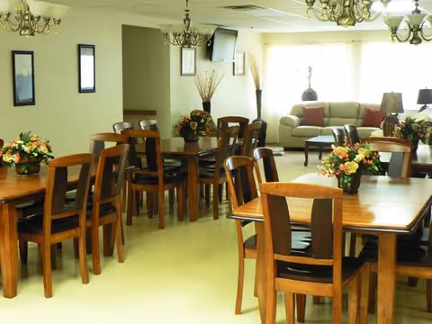 A bright and spacious dining area with multiple wooden tables and chairs arranged neatly. Each table has a floral centerpiece. In the background, there is a living room area with sofas, a coffee table, lamps, and large windows letting in natural light. The room has chandeliers hanging from the ceiling and framed pictures on the walls.