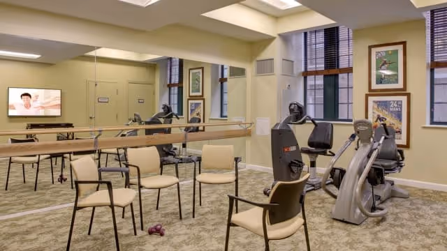 A fitness room in a senior living facility with exercise equipment including stationary bikes and a recumbent bike, several beige chairs arranged in a circle, a large mirror on one wall, framed pictures on the walls, and windows with blinds letting in natural light.