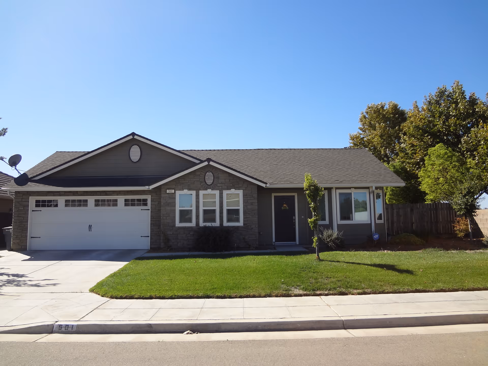 Single-story residential building with a two-car garage, stone and siding exterior, green lawn, small trees, and a clear blue sky.