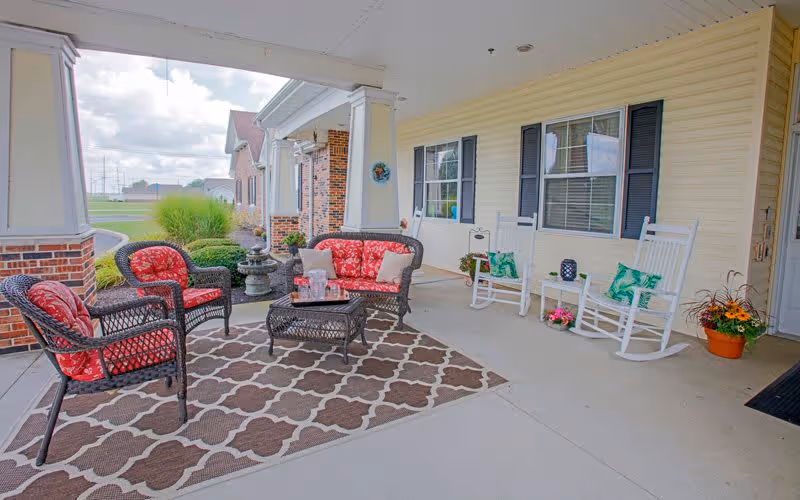 Covered outdoor patio area with wicker furniture including a loveseat and two chairs with red cushions, a coffee table on a patterned rug, two white rocking chairs with green pillows, potted plants, and a view of the lawn and neighboring buildings.