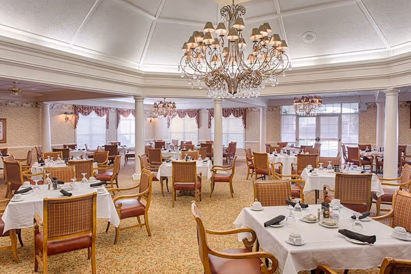 A spacious dining room in a senior living facility with multiple tables covered in white tablecloths, each set with cups, glasses, and black napkins. The room features elegant chandeliers hanging from a coffered ceiling, large windows with red and white valances, and wooden chairs with cushioned seats arranged around the tables.