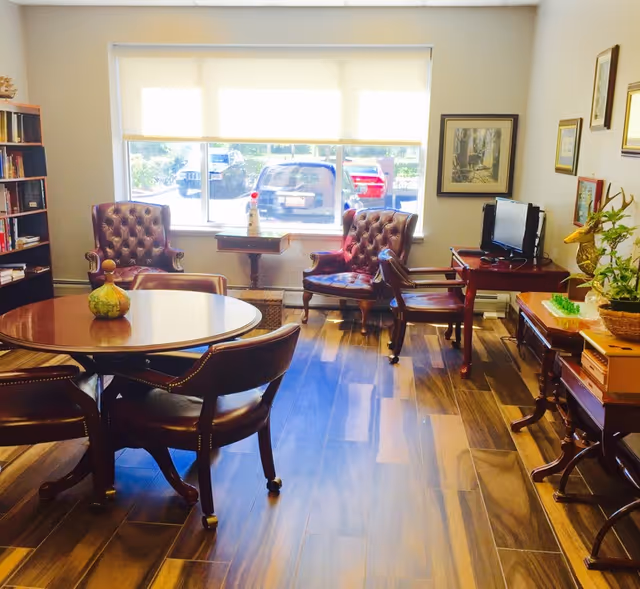A well-lit living room area with wooden flooring, featuring a round wooden table with four chairs in the foreground. Two tufted leather armchairs are placed near a large window with blinds, allowing natural light to enter. There is a small table between the armchairs and a TV on a wooden stand to the right. The room is decorated with framed pictures on the walls, a bookshelf, and various decorative items including a plant and a small sculpture.