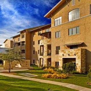 Exterior view of a multi-story senior living facility building with beige stone and stucco facade, balconies, large windows, and a well-maintained lawn with a tree and landscaping under a partly cloudy blue sky.